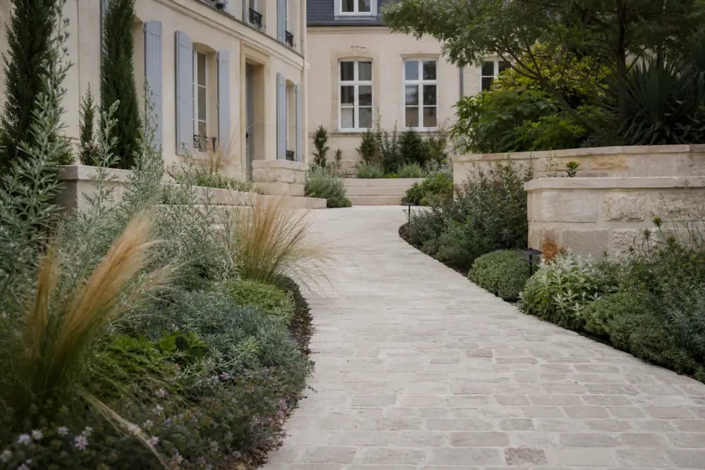 Allée de jardin en pavés courbe menant à une maison rémoise, bordée de massifs de vivaces et de graminées, lumière douce de fin de journée, sol clair rappelant la craie champenoise.