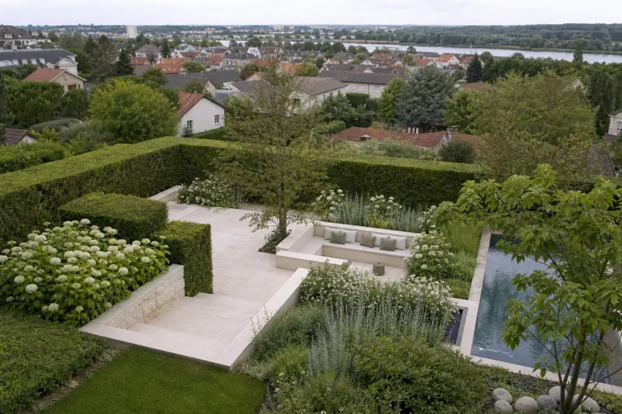 Vue aérienne basse d'un quartier pavillonnaire de la couronne de Reims : jardins verts contigus, haies taillées, la Maine en arrière-plan, lumière de fin d'après-midi.