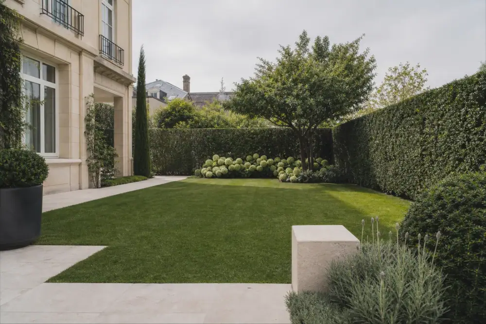 Photographie d'un jardin résidentiel soigné à Reims, pelouse fraîchement tondue et haies taillées nettes, lumière douce de fin d'après-midi, un propriétaire détendu profitant de son extérieur en arrière-plan.